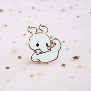 Puede incluir: Un pin de esmalte azul claro con forma de perro fantasma de dibujos animados con un contorno dorado. El pin tiene una pequeña nariz negra, una boca rosa y una nariz dorada. El pin está sobre un fondo rosa con estrellas doradas.