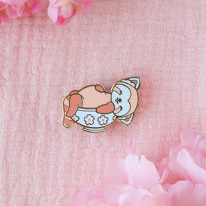 May include: A blue and pink enamel pin of a red panda sleeping in a bowl with a floral pattern.