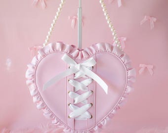 May include: A pink heart-shaped handbag with white lace trim and a white ribbon corset detail. The bag has a pearl chain strap.