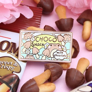 Puede incluir: Un pin de esmalte dorado con una rana de dibujos animados sosteniendo una caja de bocadillos de chocolate. La caja dice "Choco Snack" y "chocolate con leche". El pin está rodeado de palitos de galletas cubiertos de chocolate.