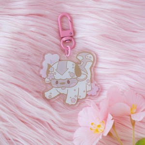 May include: A pink and white acrylic keychain featuring a cartoon Appa from Avatar: The Last Airbender. Appa is surrounded by pink cherry blossoms. The keychain has a pink metal clasp.