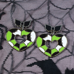 May include: A pair of black and green heart-shaped earrings with a white patchwork design. The earrings have a black bow at the top.