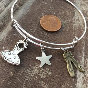 May include: Silver adjustable bracelet with three charms: a ballet dress, a star, and a pair of ballet shoes. A penny is next to the bracelet. The bracelet is on a wooden surface.
