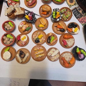 May include: A collection of miniature food items arranged on small round plates and in tiny bowls. The assortment includes various sculpted dishes like tacos, tamales, rice, beans, and vegetables, featuring vibrant colours and detailed textures.