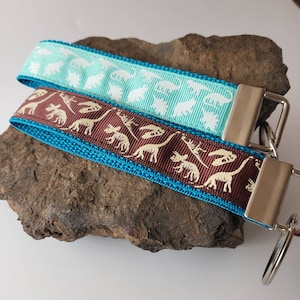 May include: Two keychains featuring dinosaur skeleton designs. One is turquoise blue with white skeletons, the other is brown with cream skeletons. Both have silver key rings.