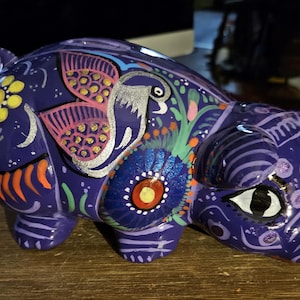 Op de afbeelding: Een paarse keramische spaarpot met kleurrijke beschilderde details, waaronder bloemen, vogels en swirls. De spaarpot heeft een groot oog met een zwarte pupil en witte highlights.