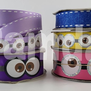 May include: Spools of decorative ribbon in various colors, including purple, pink, yellow, and blue. Some ribbons feature a repeating eye design with brown irises and silver glitter accents. The top ribbons have a stitched edge.