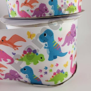 Könnte beinhalten: Weiße Bandspulen mit einem sich wiederholenden Muster aus bunten Zeichentrick-Dinosauriern, Schmetterlingen und Fußabdrücken. Die Dinosaurier sind in Blau, Grün, Rosa, Lila und Orange gehalten. Geeignet für Bastelarbeiten, Geschenkverpackungen und Partydekorationen.