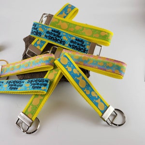Op de afbeelding: Een set van vijf gele en blauwe sleutelhangers met een vispatroon en de tekst "gemakkelijk afgeleid door snacks".