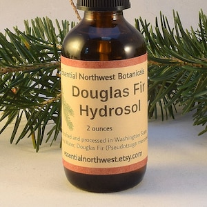 May include: A brown glass bottle with a black spray top, labelled "Essential Northwest Botanicals Douglas Fir Hydrosol 2 ounces Crafted and processed in Washington State Water, Douglas Fir (Pseudotsuga menziesii) essentialnorthwest.etsy.com"