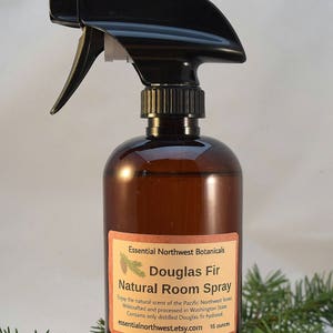May include: A brown glass bottle with a black spray nozzle filled with Douglas Fir Natural Room Spray. The bottle has a label with the text "Essential Northwest Botanicals Douglas Fir Natural Room Spray" and the text "Enjoy the natural scent of the Pacific Northwest forest Wildcrafted and processed in Washington State Contains only distilled Douglas fir hydrosol essentialnorthwest.etsy.com 16 ounces". The bottle is surrounded by green fir branches.