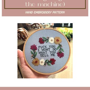 Peut inclure: Motif de broderie à la main avec le texte "FUCK YOU I WON'T DO WHAT YOU TELL ME" entouré de fleurs brodées rouges, blanches et dorées. Le texte "female rage (against the machine)" est également présent.