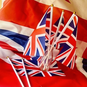 Puede incluir: Tres pequeñas banderas de la Union Jack en palos blancos con puntas doradas, mostradas sobre un fondo de tela rojo, blanco y azul.