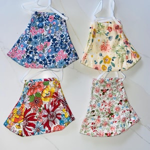 May include: Four floral face masks with white straps are displayed. Each mask features a different colorful floral pattern, including blue, pink, red, yellow, and green. The masks are hanging on white hangers, showcasing their design and shape.