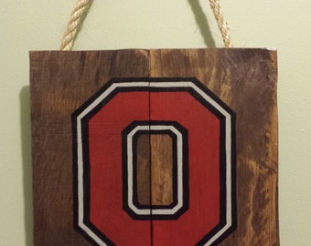 May include: A wooden sign with a red and white "O" painted on it. The sign is hanging from a rope and is attached to a hook on the wall.