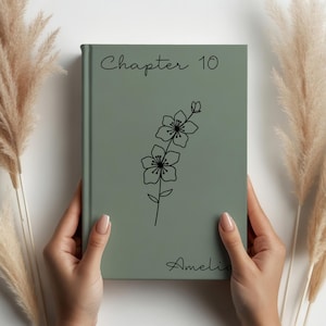以下が含まれることがあります： 「Chapter 10」のテキストと「Amelia」の名前が書かれたセージグリーンの本。黒い花のイラストが表紙の中央にあります。本は、乾燥した観賞用の草がある白い背景の前に保持されています。