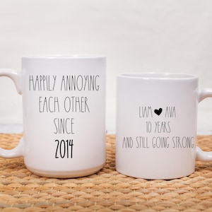 May include: Two white ceramic mugs with black text. The mug on the left says "Happily annoying each other since 2014". The mug on the right says "Liam Ava 10 years and still going strong".