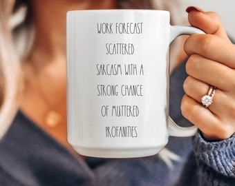 May include: White ceramic mug with the text "WORK FORECAST SCATTERED SARCASM WITH A STRONG CHANCE OF MUTTERED PROFANITIES" in black. The mug has a large handle. The mug is held by a person.