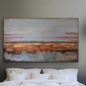 May include: A bedroom with a large abstract landscape painting above a bed with white bedding. The painting depicts a blue sky, a golden-brown horizon, and a water reflection. Two woven pendant lights hang from the ceiling.