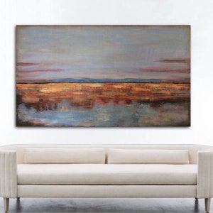 May include: Abstract landscape painting with a muted colour palette. The artwork features a sky of grey and pink hues above a horizon of orange, brown, and blue tones, with reflections in the water below. A cream-coloured sofa is below the painting.