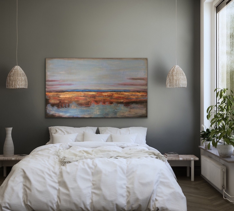 May include: A bedroom with a large abstract landscape painting above a bed with white bedding. The painting depicts a blue sky, a golden-brown horizon, and a water reflection. Two woven pendant lights hang from the ceiling.
