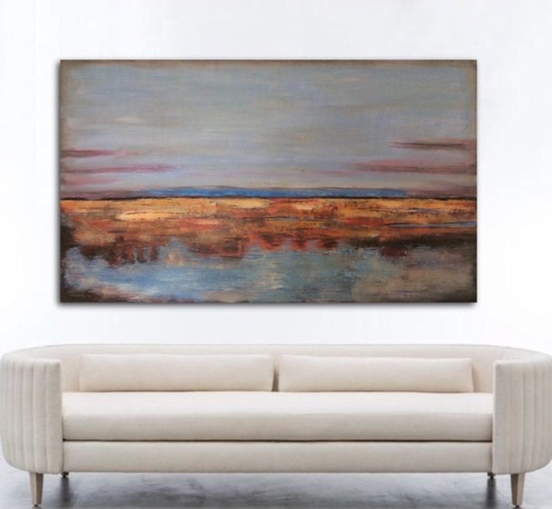 May include: Abstract landscape painting with a muted colour palette. The artwork features a sky of grey and pink hues above a horizon of orange, brown, and blue tones, with reflections in the water below. A cream-coloured sofa is below the painting.