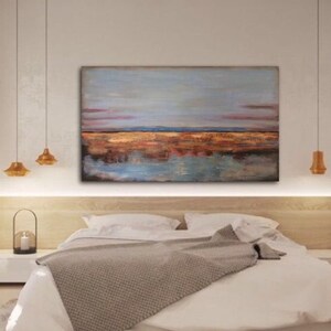 May include: An abstract landscape painting in shades of blue, brown, and orange. The artwork shows a horizon line and its reflection in the water. The painting hangs above a bed with white bedding and a grey blanket. The artwork is a decorative piece.