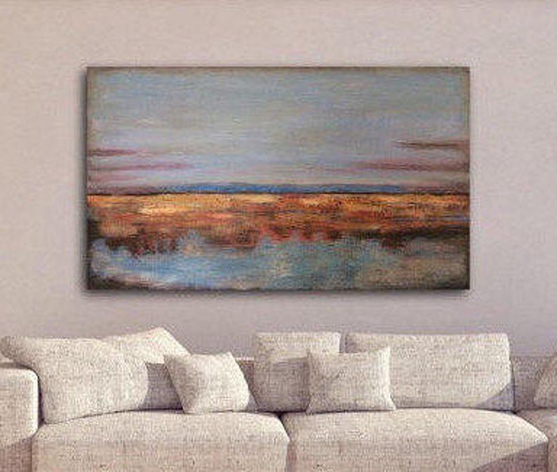 May include: An oil painting of a landscape with a reflective body of water. The artwork shows a sky with blue and pink hues, a golden-brown field, and a mirrored reflection in the water. The painting is displayed above a beige sofa.