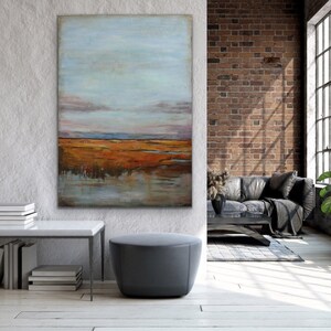 May include: A large abstract painting of a landscape with a blue sky, orange and brown fields, and a reflection of the sky in the water. The painting is hanging on a white wall in a modern living room.