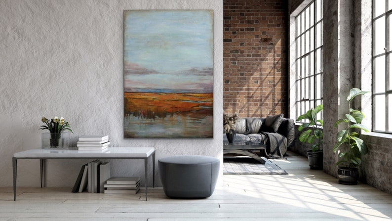 May include: A large abstract painting of a landscape with a blue sky, orange and brown fields, and a reflection of the sky in the water. The painting is hanging on a white wall in a modern living room.