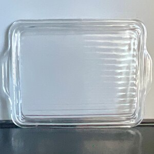May include: A clear, rectangular glass baking dish with rounded corners and two small handles on the sides. The dish has a ribbed texture on the bottom and is designed for oven use. The glass is transparent, allowing visibility of the contents.