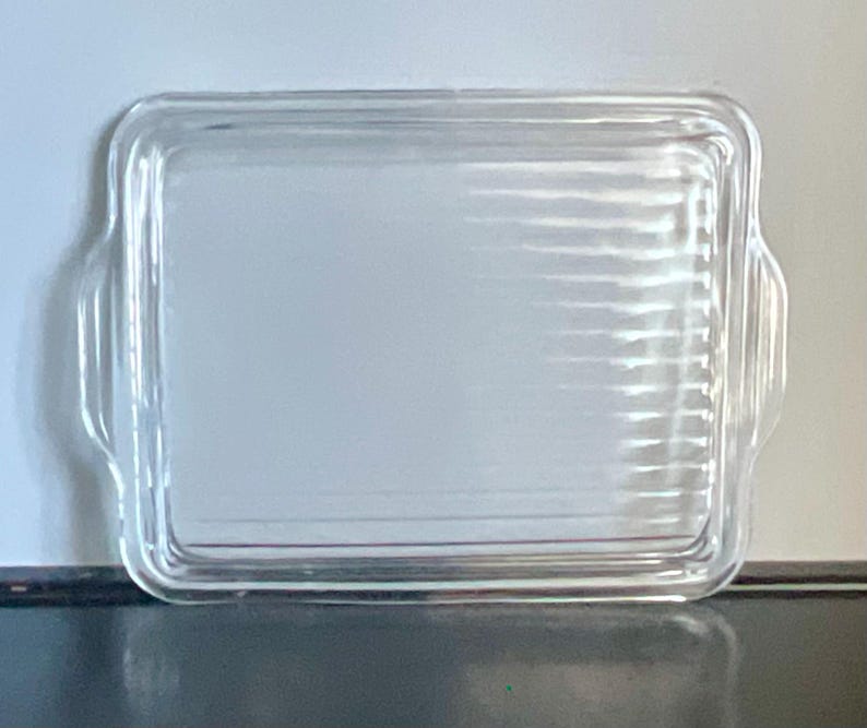 May include: A clear, rectangular glass baking dish with rounded corners and two small handles on the sides. The dish has a ribbed texture on the bottom and is designed for oven use. The glass is transparent, allowing visibility of the contents.