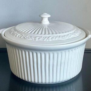 May include: A white ceramic casserole dish with a matching lid. The round vessel features vertical ribbed detailing along the exterior walls and two small side handles. The lid has a central knob and a decorative embossed scroll pattern around the edge.