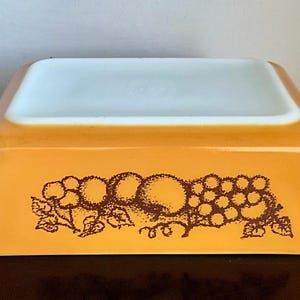 May include: An orange Pyrex baking dish with a white interior. The exterior features a brown fruit design, including grapes and other fruits. The dish has a rectangular shape with slightly curved sides and small handles.