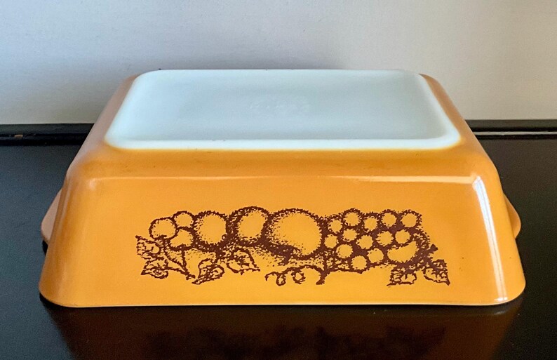 May include: An orange Pyrex baking dish with a white interior. The exterior features a brown fruit design, including grapes and other fruits. The dish has a rectangular shape with slightly curved sides and small handles.
