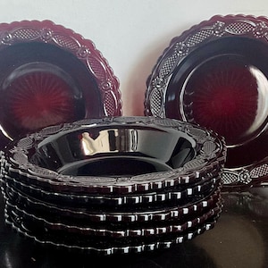 Puede incluir: Un juego de platos y cuencos de cristal rojo oscuro con borde festoneado y un patrón texturizado en relieve a lo largo del borde. La colección incluye dos platos verticales colocados detrás de una pila de cinco cuencos a juego, en tono rubí.