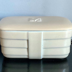 May include: A rectangular, cream-colored ceramic box with a matching lid. The box features horizontal ribbed detailing and a vertical band on the front. A small, stylized design is centered on the lid.