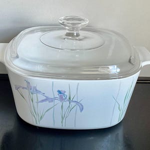 May include: White casserole dish with a clear glass lid and handle. The dish features a floral design with purple and blue irises and green stems. The dish is square with rounded corners.