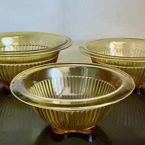 May include: Three amber-colored glass bowls with a ribbed exterior and a smooth, rounded rim. The bowls are of varying sizes and sit on a dark surface. The bowls are a vintage kitchenware set.