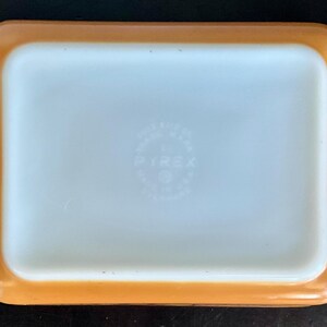 May include: Rectangular Pyrex baking dish with a light orange exterior and a white interior. The dish has rounded corners and small handles on each side. The Pyrex logo is visible in the center of the dish.