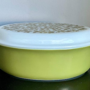 May include: A vintage, lime green casserole dish with a white lid. The lid is decorated with a gold floral pattern. The dish has a rounded shape with handles on either side. A classic piece of kitchenware.