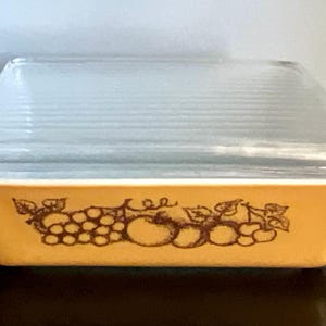 May include: Rectangular, amber-colored Pyrex baking dish with a clear, ribbed glass lid. The dish features a brown fruit design around the base. A vintage kitchenware item.