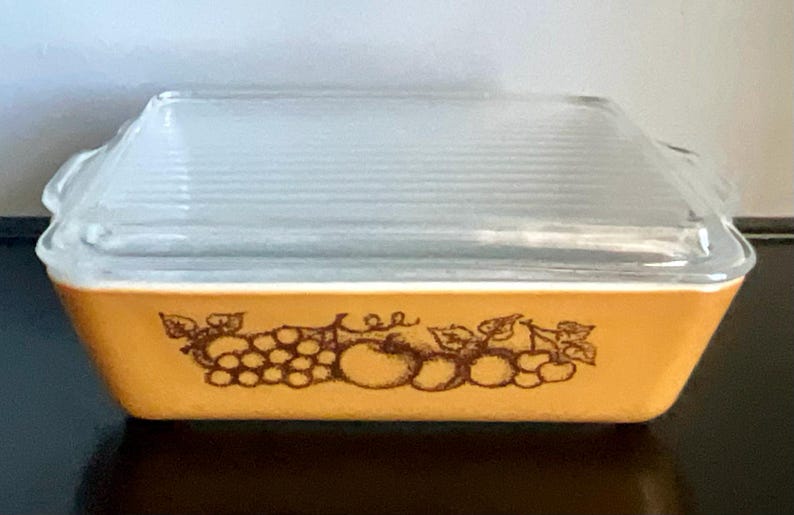 May include: Rectangular, amber-colored Pyrex baking dish with a clear, ribbed glass lid. The dish features a brown fruit design around the base. A vintage kitchenware item.