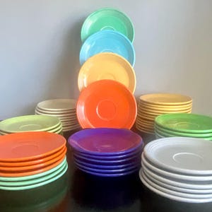 May include: A collection of colorful ceramic plates in various shades, including orange, green, blue, yellow, purple, and white. The plates are stacked in neat piles and arranged to showcase their vibrant hues. The plates are a classic design.