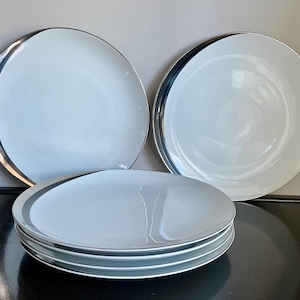 May include: Set of four white ceramic dinner plates with a silver rim. The plates are stacked on top of each other.