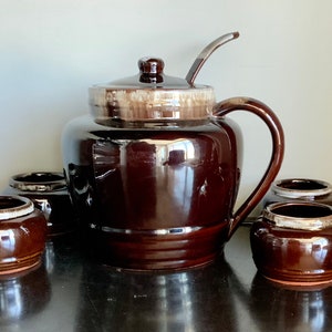 May include: A brown ceramic punch bowl with a lid and ladle, and six matching cups. The bowl has a white glaze on the top edge and the cups have a white band around the top.