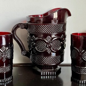 May include: A dark red glass pitcher and four matching tumblers featuring a raised diamond pattern and circular motifs. The pitcher has a curved handle and a wide pouring spout, while each glass has a cylindrical shape with a textured base.