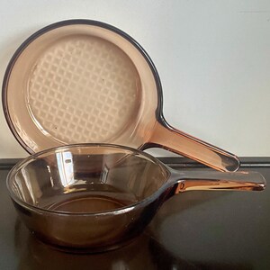 May include: Two amber-colored glass skillets with integrated handles. The larger skillet features a textured grid pattern on the interior base, while the smaller skillet has a smooth, transparent interior surface and a deeper, rounded bowl shape.