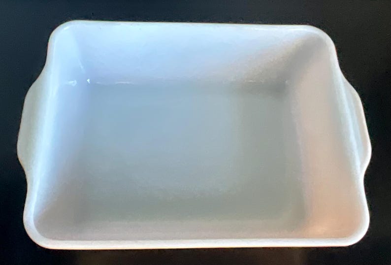 May include: A rectangular, white baking dish with rounded corners and two small handles on the sides. The dish has a smooth, glossy surface and is empty. The overall shape is a classic design for baking and serving.