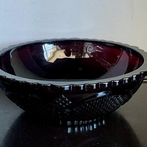 May include: A deep red glass bowl featuring a scalloped rim and a textured exterior pattern consisting of diamond shapes and cross-hatching. The bowl has a smooth, reflective interior surface and a wide, circular opening with a thick, molded edge.